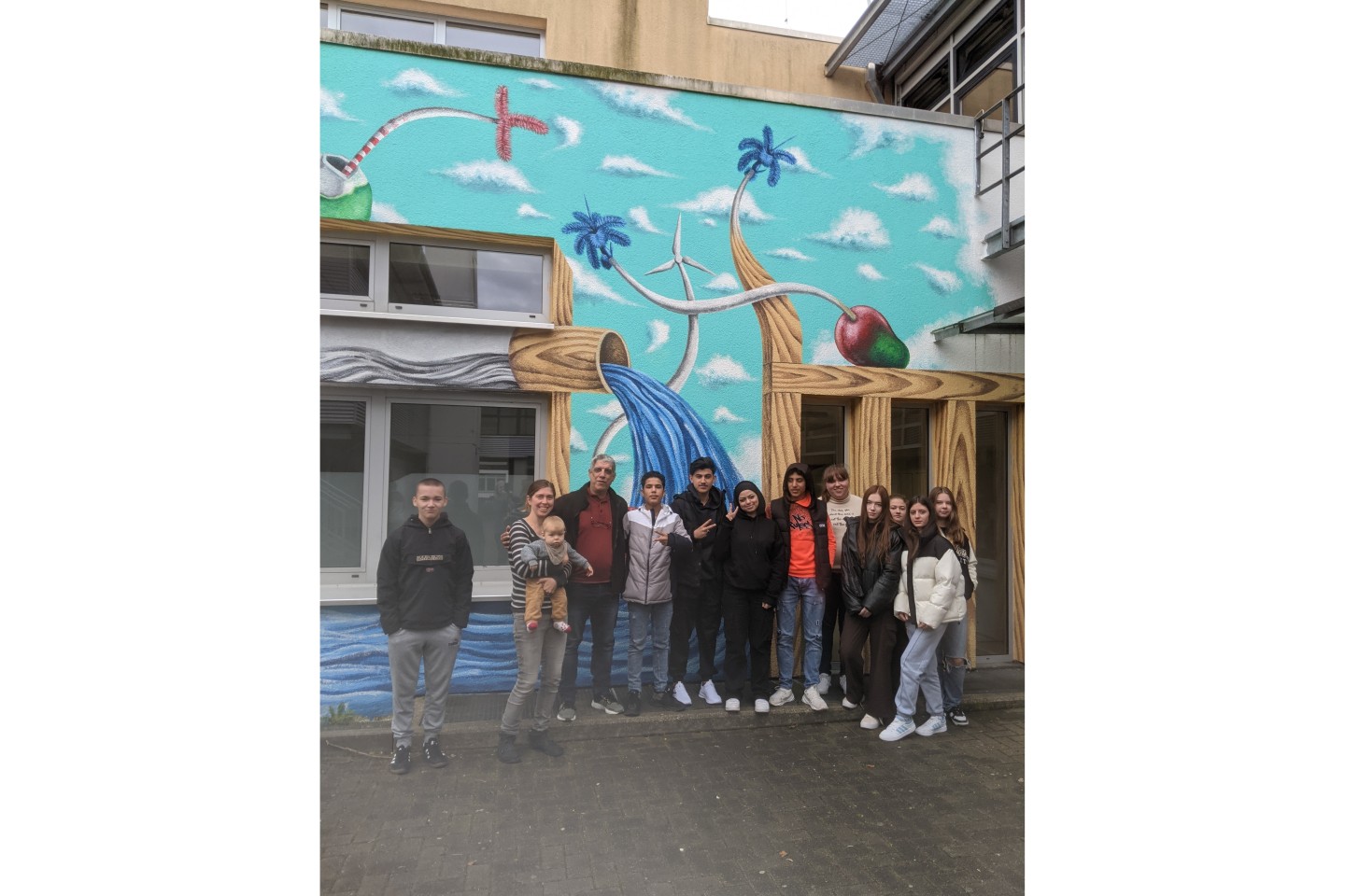 Fotounterschrift: Abel Morejón Galá (3.v.l.) zusammen mit Kunstkollegin Lina Riebler (2. v.l.) und den Schüler:innen der Klasse 9e vor dem Wandgemälde an der Geschwister-Scholl-Straße